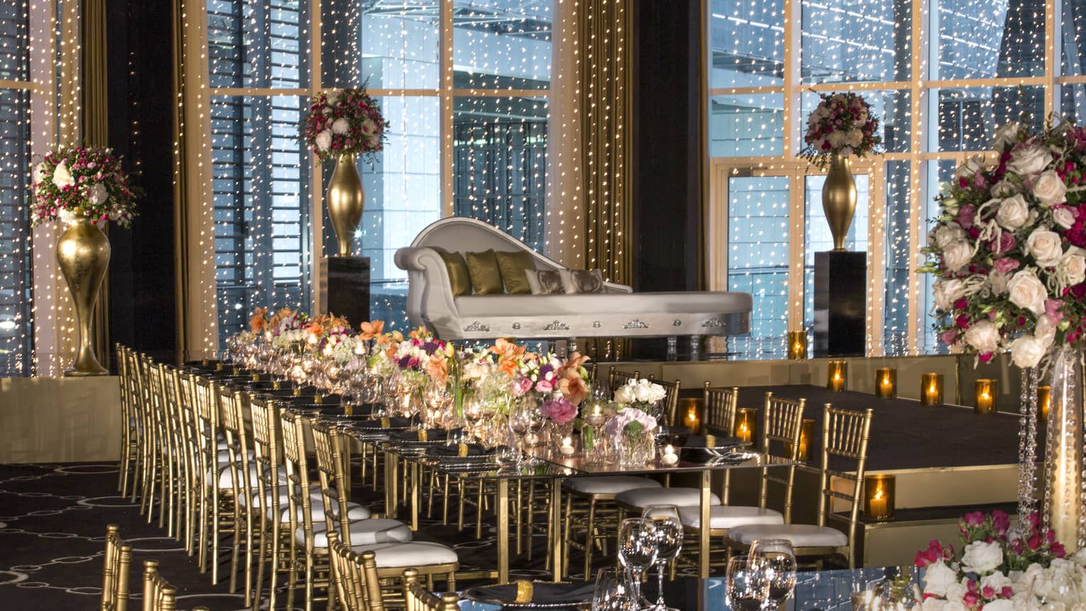 Wedding banquet table with flower arrangements, gold chairs and chaise below window with string lights 
