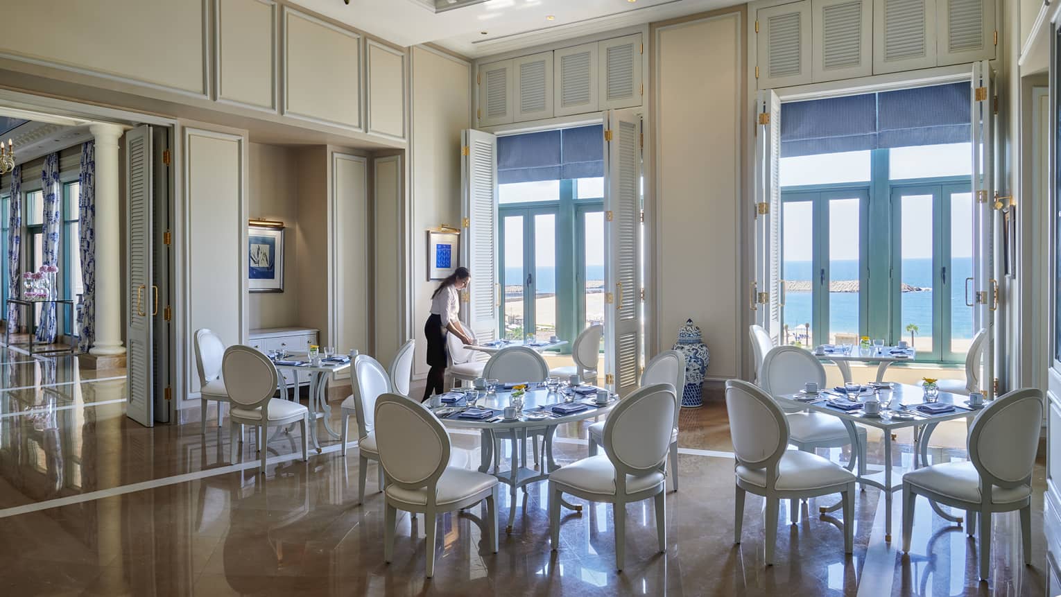 Server sets white table at Kala restaurant, white shutters with blue blinds lead to ocean views 