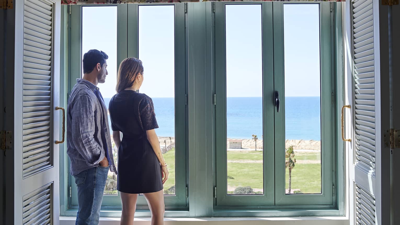 Couple stand at large rustic window overlooking Four Seasons Hotel Alexandria lawns, waterfront 
