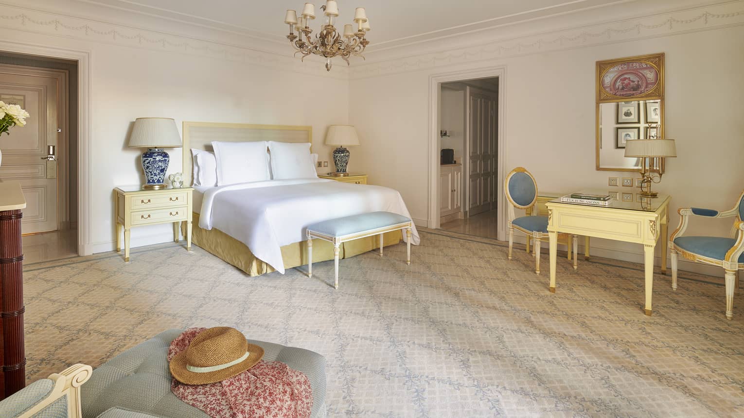 Bedroom with white king bed, light blue bench, two blue chairs and beige wooden table, chandelier