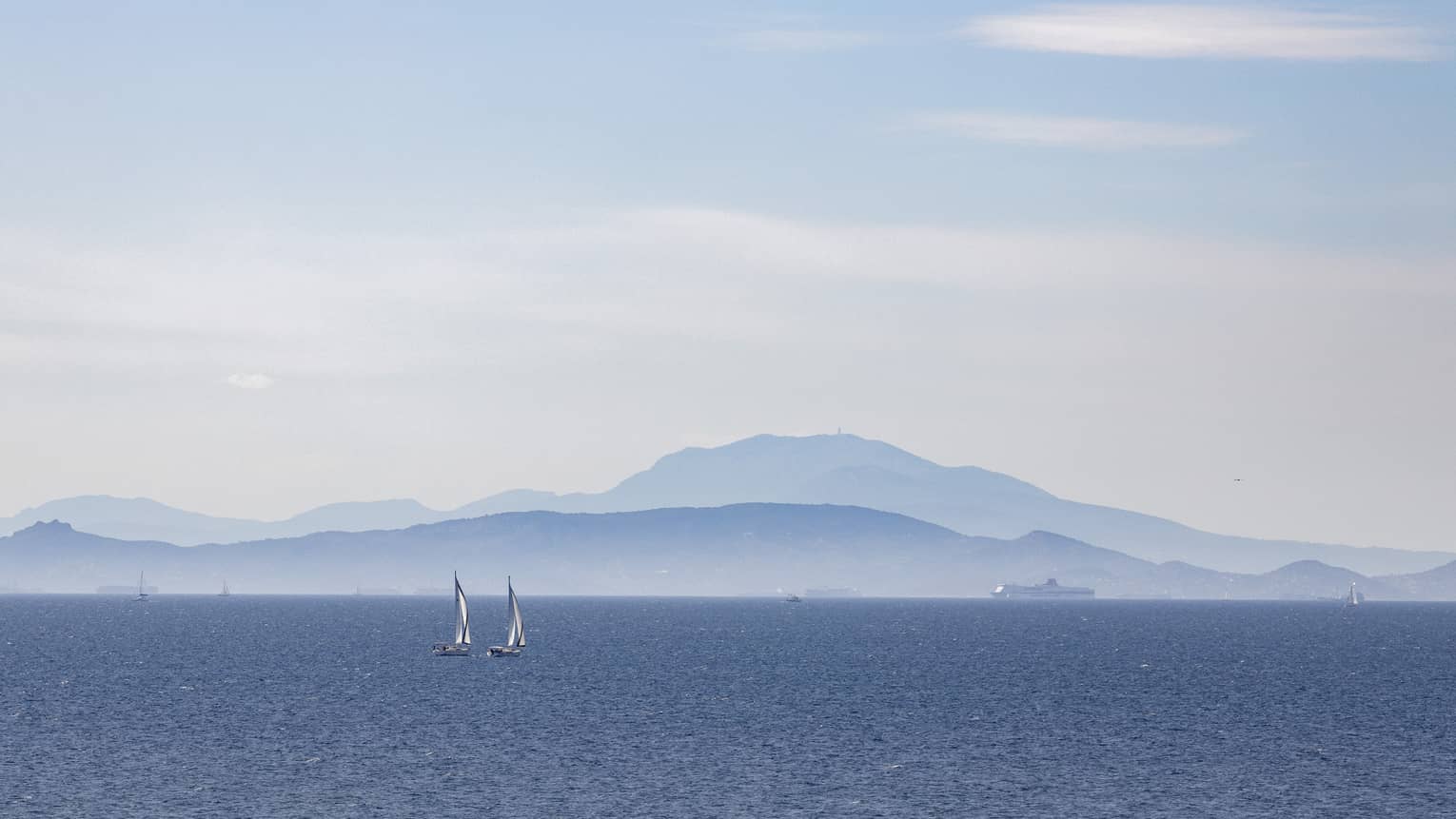 Two sailboats on blue ocean with blue-grey mountain range in the background 