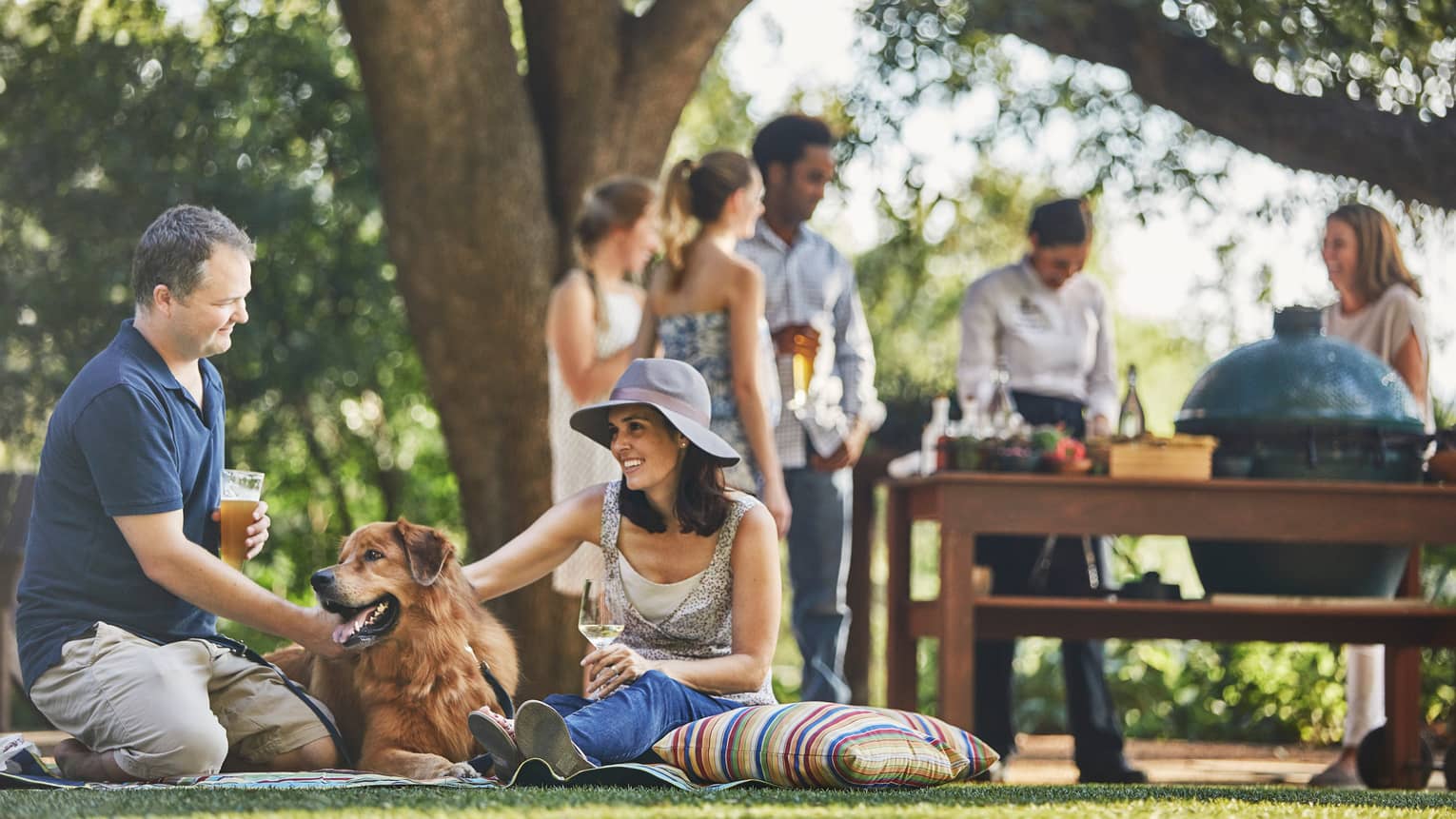 Man with beer, woman with wine sit on lawn, pet large dog, people around barbecue in background