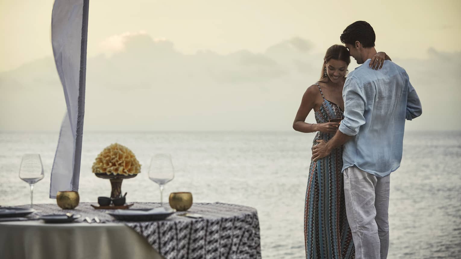 Couple embraces beside private dining table overlooking ocean at sunset