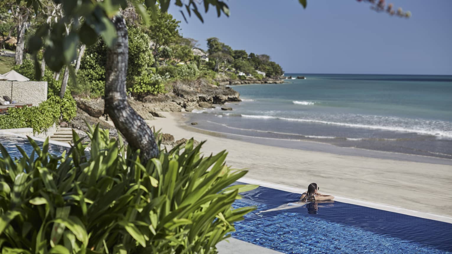 View from tropical plants looking down at swimming pool where woman rests head on ledge, looks out at Sundara beach