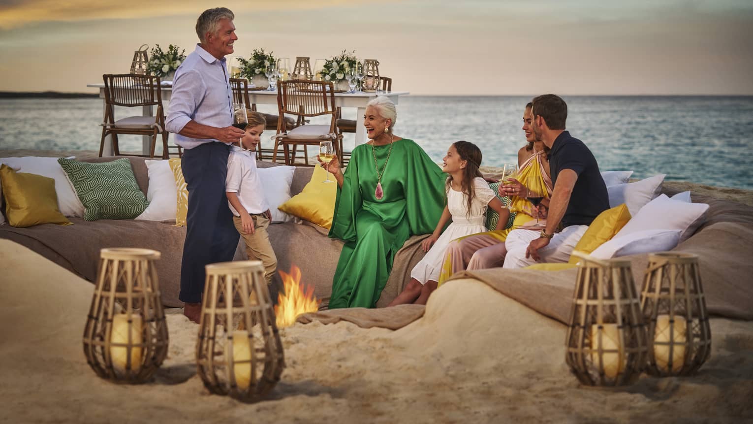 Family sitting in the sand around a bonfire with a dining table behind them.