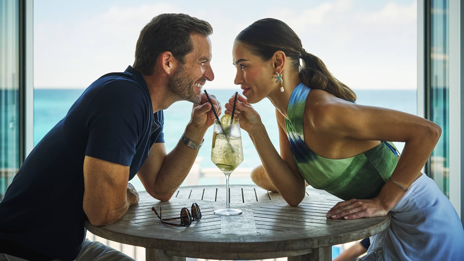 Man and woman sitting at a dinner table with a view of the ocean, sharing a drink.