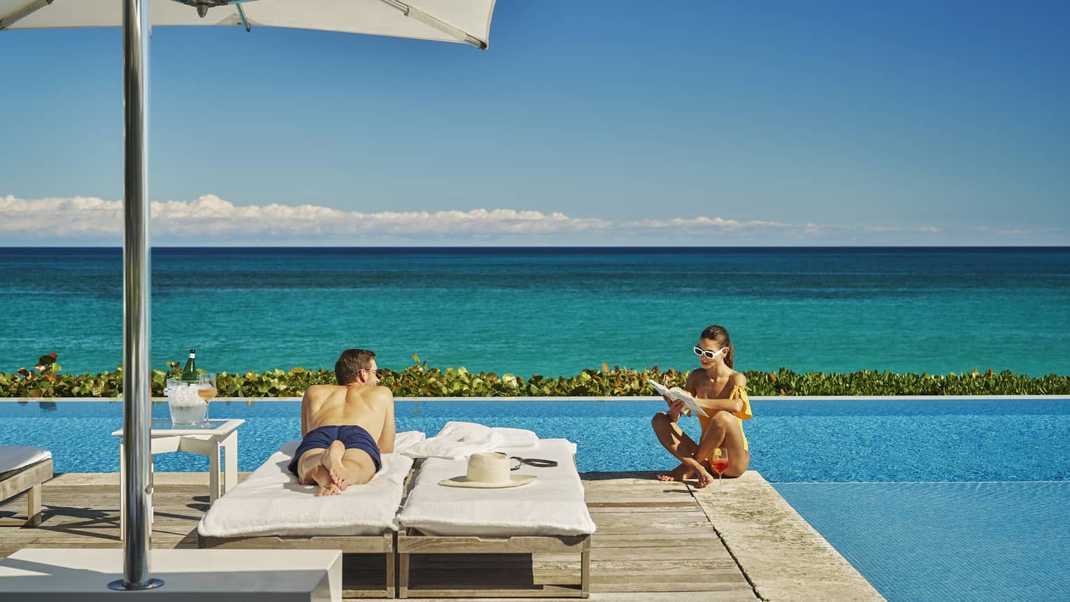 Man laying in a lounge chair by the pool while woman sits on the edge of the pool.