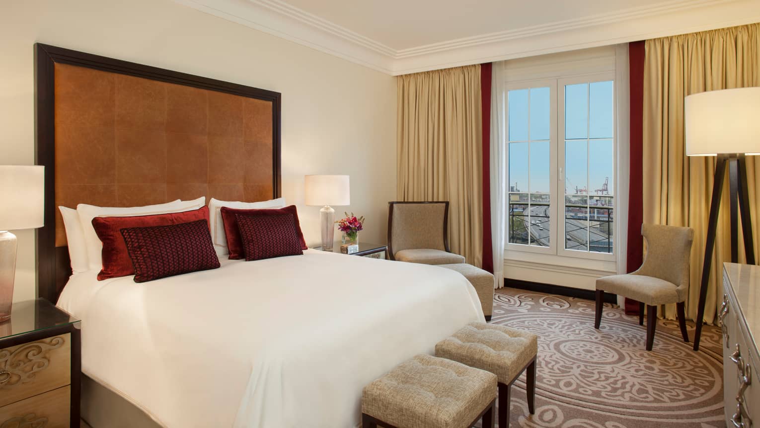 City-View One Bedroom Suite bed with tall padded leather headboard, small red accent pillows, two chairs by window