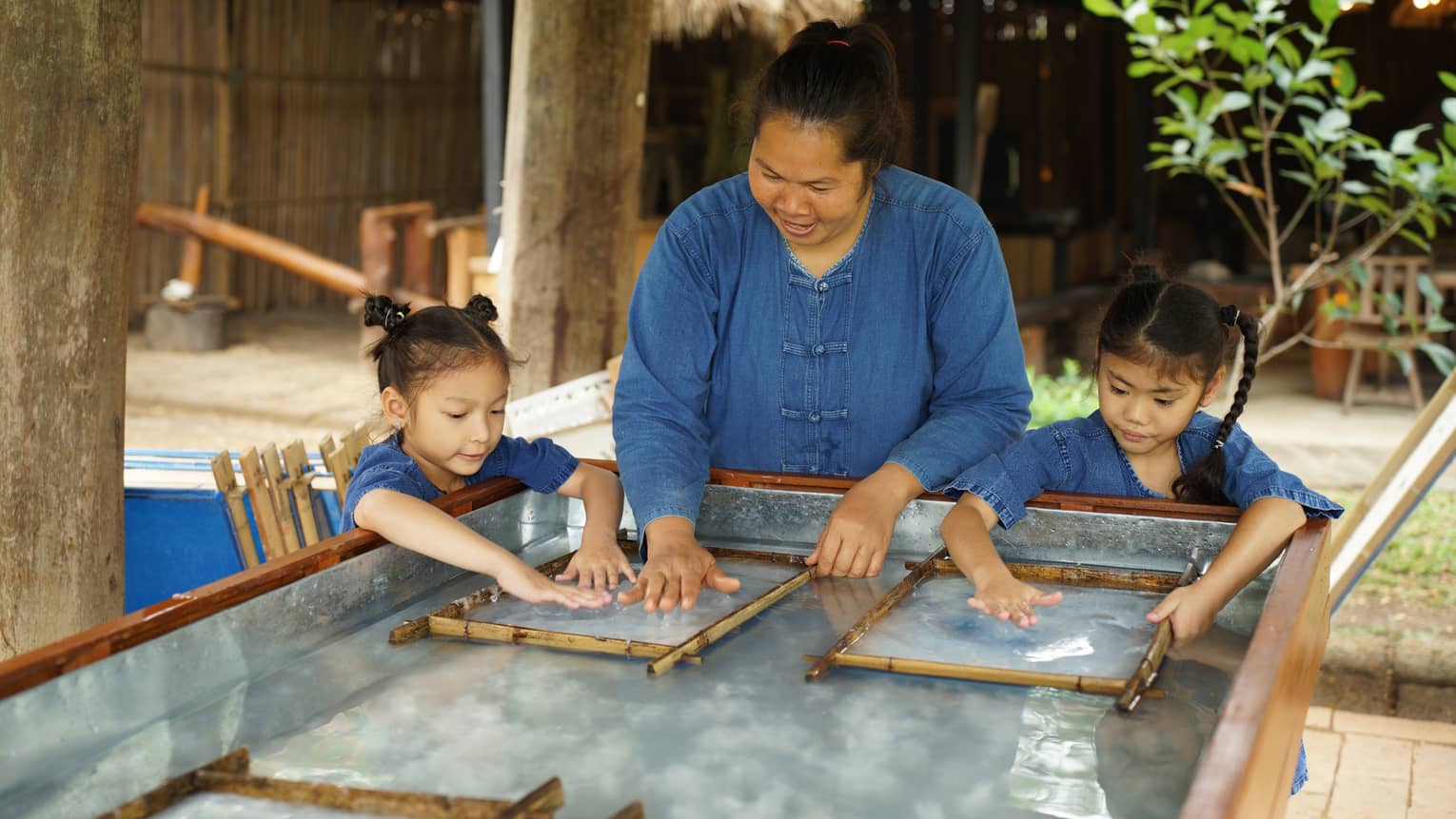 An adult stands between two children, running their hands along a clear, wet substance enclosed in two thin bamboo frames.