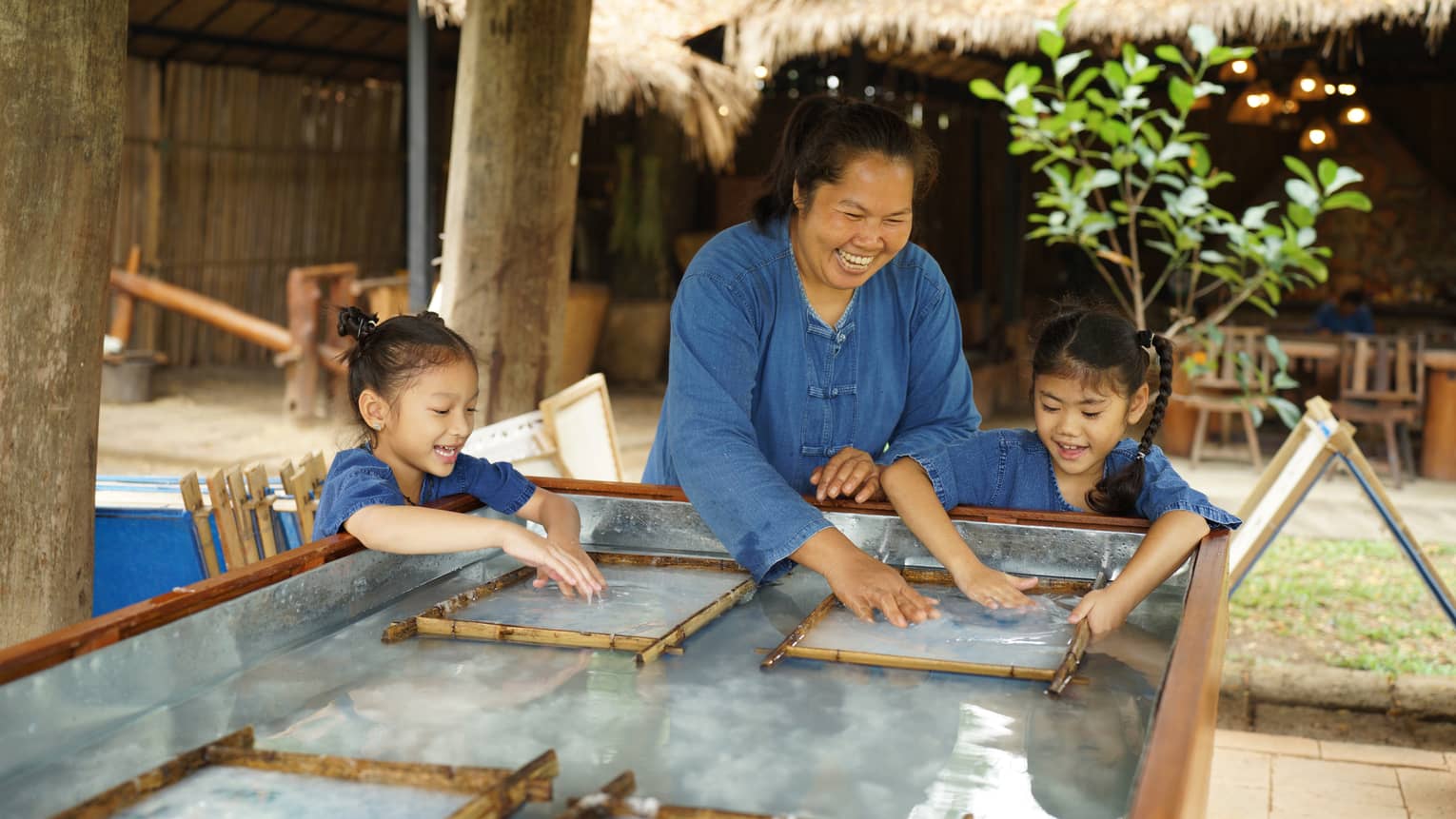 An adult and two children laugh as they smooth over by hand a clear, wet substance held within two thin bamboo frames.