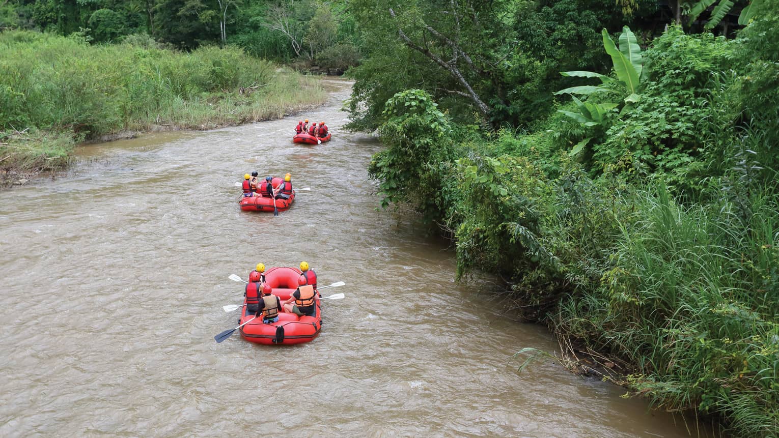 Rear view of three red inflatable rafts, five helmeted riders in each, coasting down a winding river flanked by lush foliage.