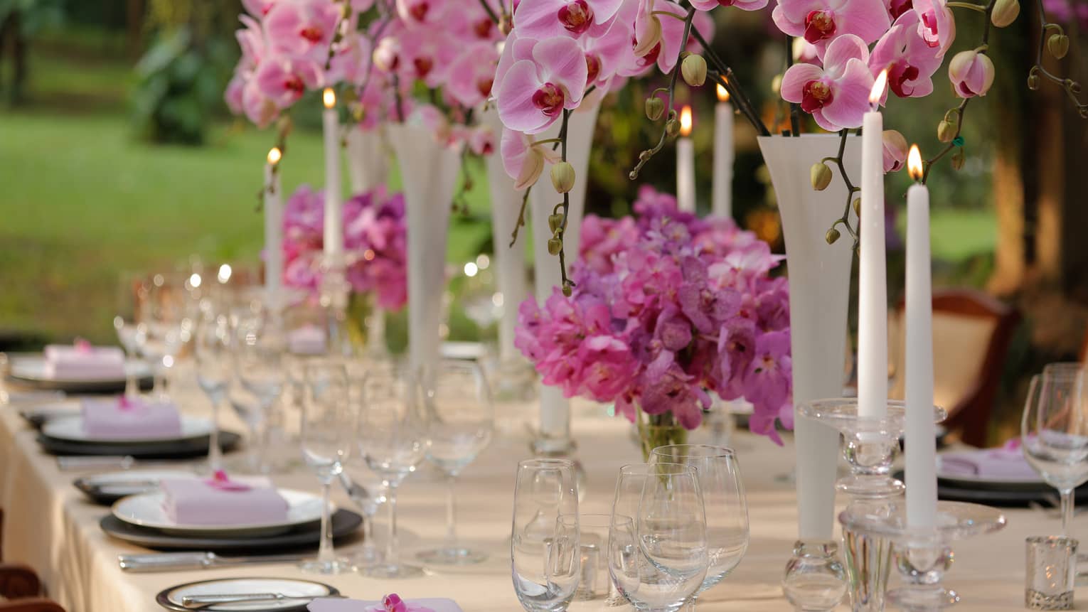 Outdoor dining table with pink flowers in vases, tall white candles 
