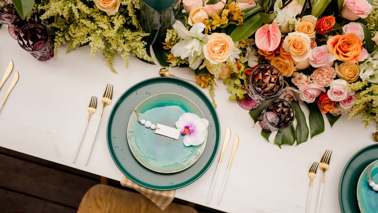 Two teal plates are topped with a white-and-pink flower, with gold cutlery on either side with a large centerpiece featuring various pink and peach flowers along with greenery all set on a white tablecloth
