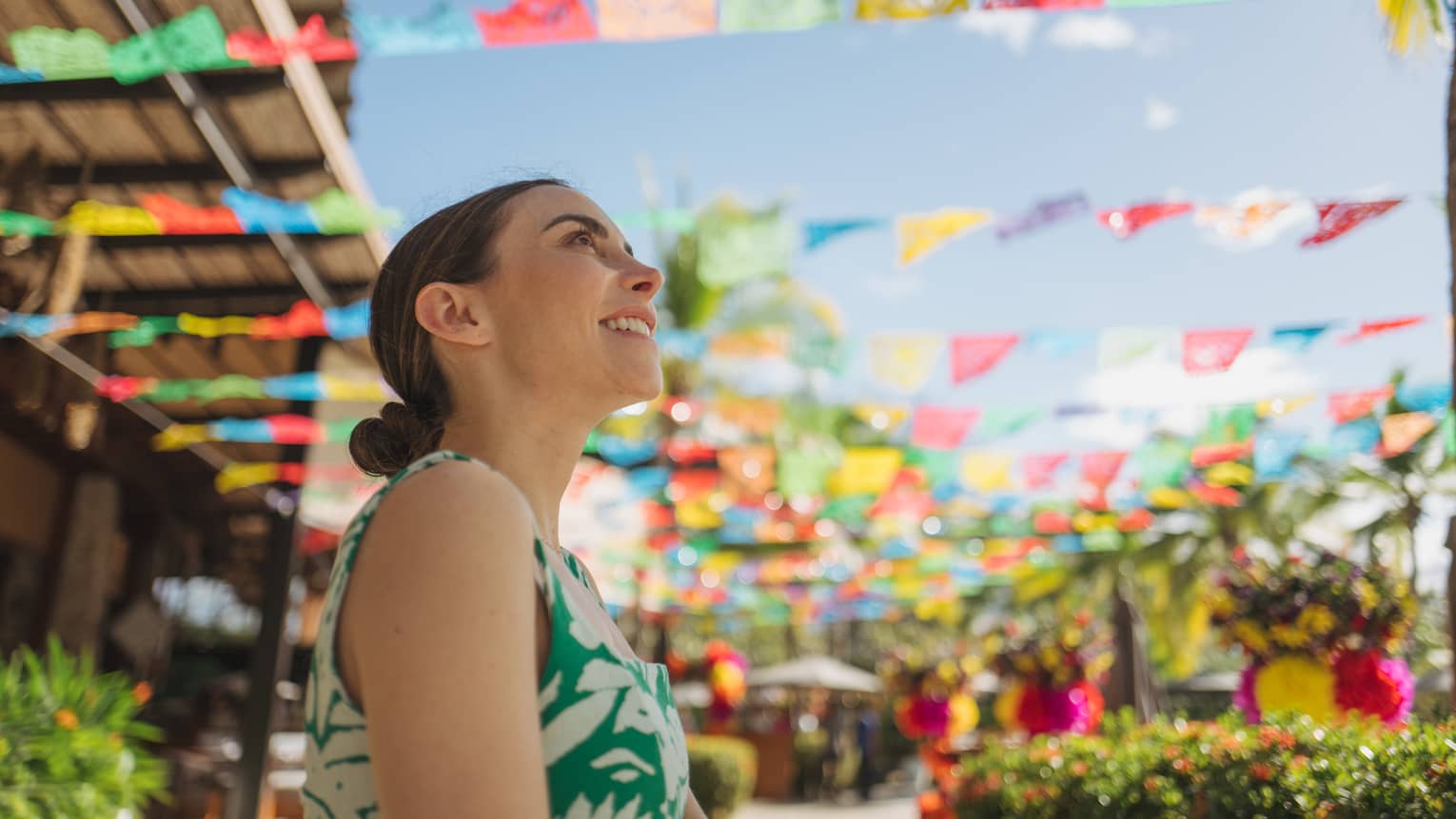 Woman with her hair pulled back in a low bun and wearing a green-and-white patterned dress stands beneath rows of colored squares hanging in an outdoor market
