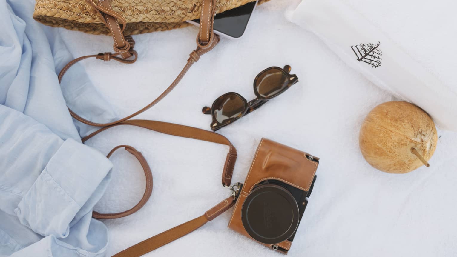 A light-blue overshirt, straw basket with brown leather straps, sunglasses, a camera in a brown leather case, a white Four Seasons towel and a coconut with a straw sticking out of it lay on top of a crisp white blanket