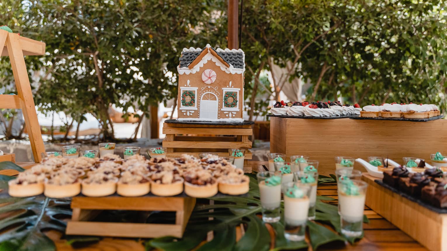 Wooden table set with holiday-themed desserts including cookies, tarts, merengues, custard-in-a-glass and a gingerbread house, all placed on pallet-style stands of varying heights