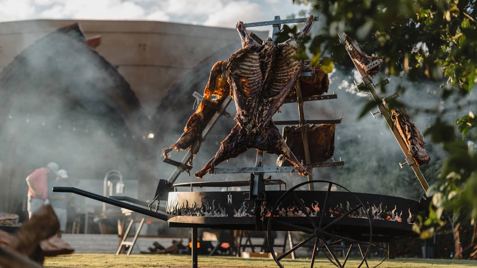 Six large piece of beef hang above a large outdoor asado grill as smoke billows in the distance