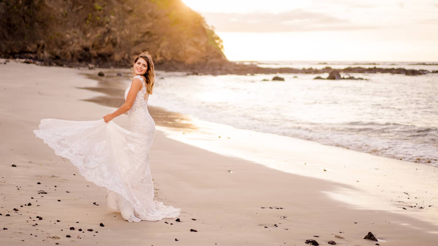 Bride in lace mermaid-style wedding gown holds up her train as she walks along the beach at sunset