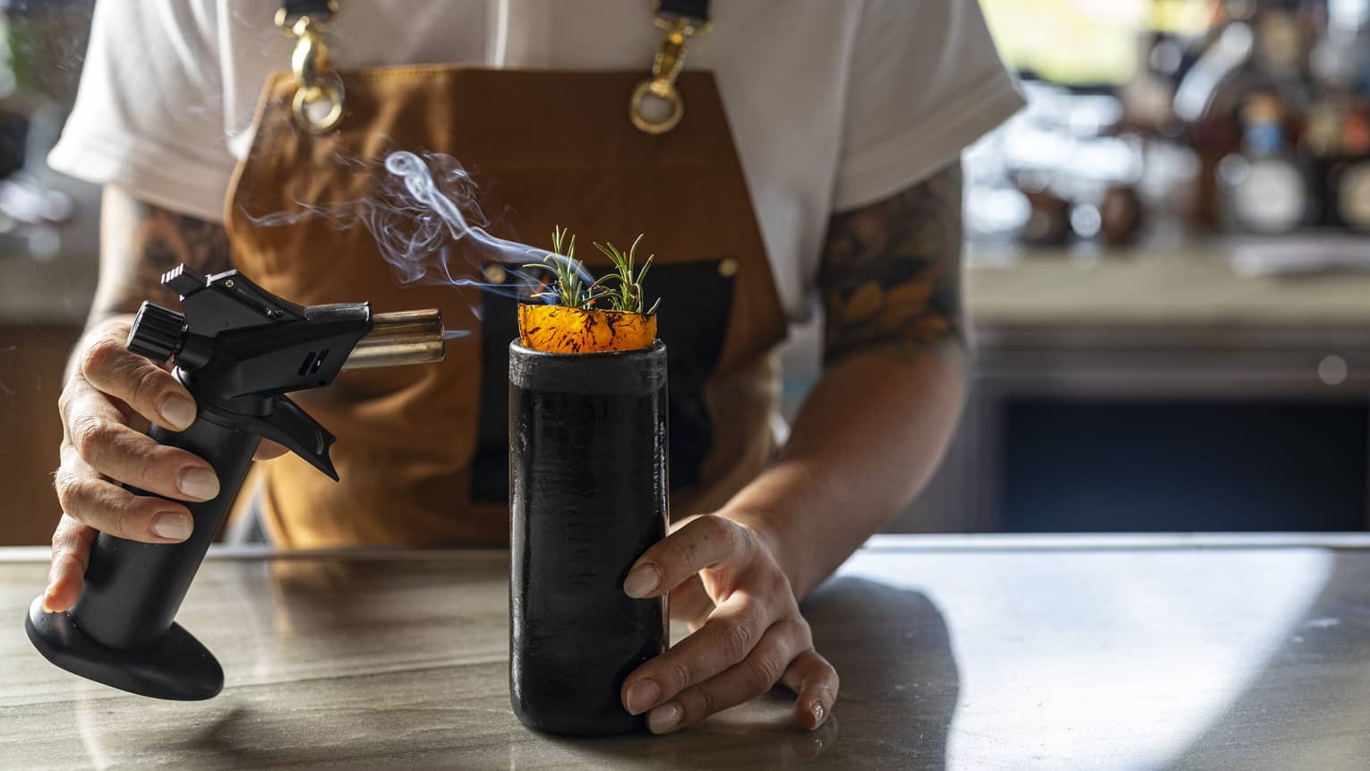 A bartender wearing a brown canvas apron holds a small blow torch in one had as they light the garnish coming out of the top of a tall, dark cocktail glass