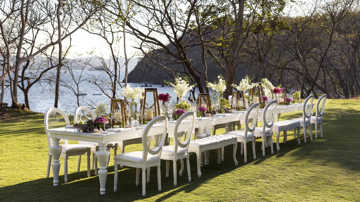 White dining table with fresh flowers, lanterns on sunny lawn overlooking ocean