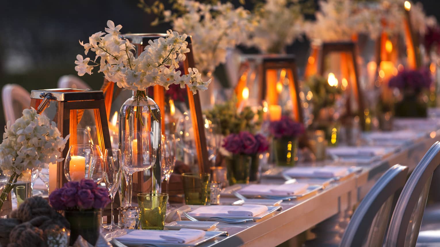 Close-up of candles in glowing lanterns, vases with flowers on long dining table at night