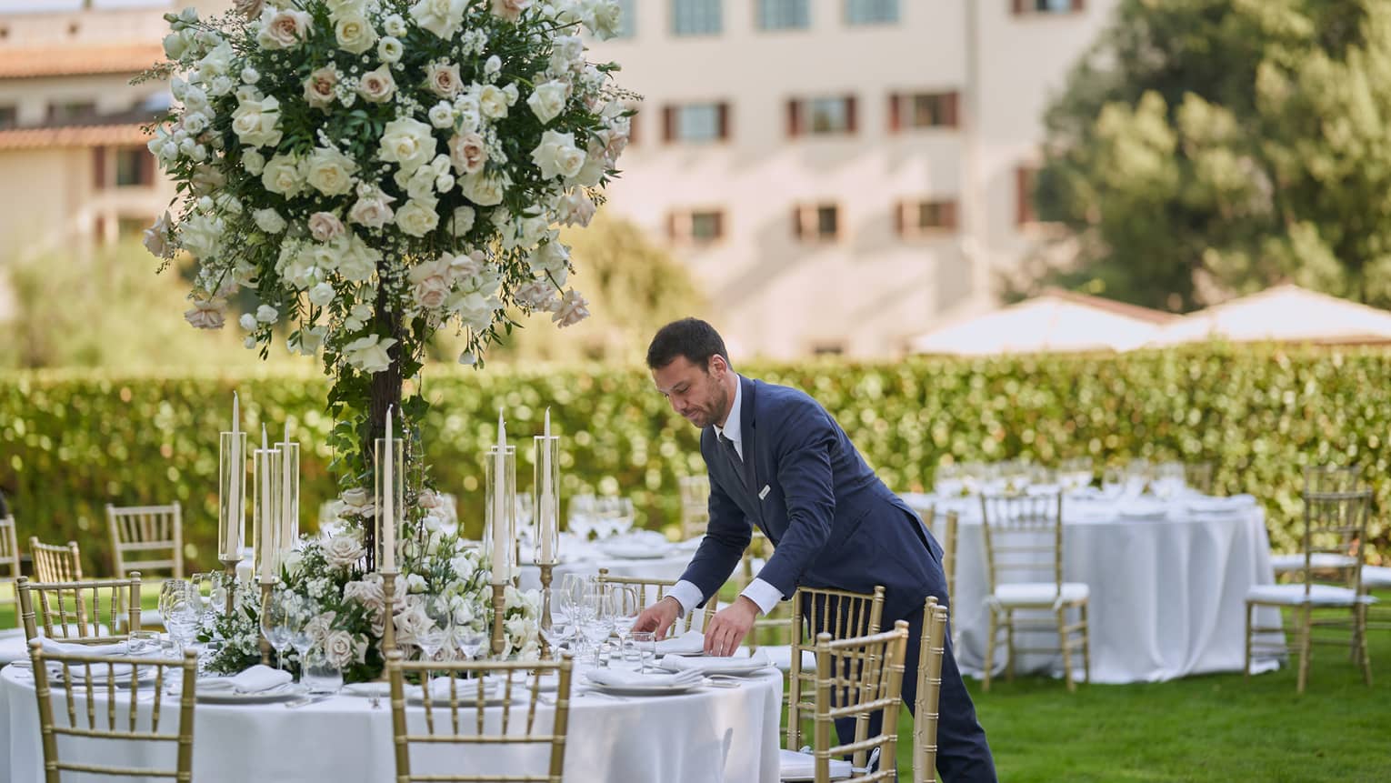 Man in suit sets garden wedding reception tables with large white topiary and hotel in backdrop