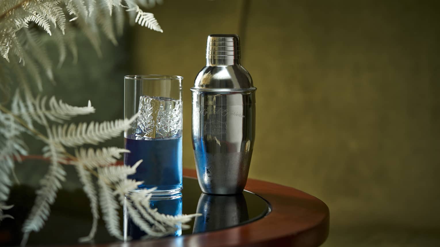 Blue cocktail in crystal highball glass beside chrome cocktail shaker and fern