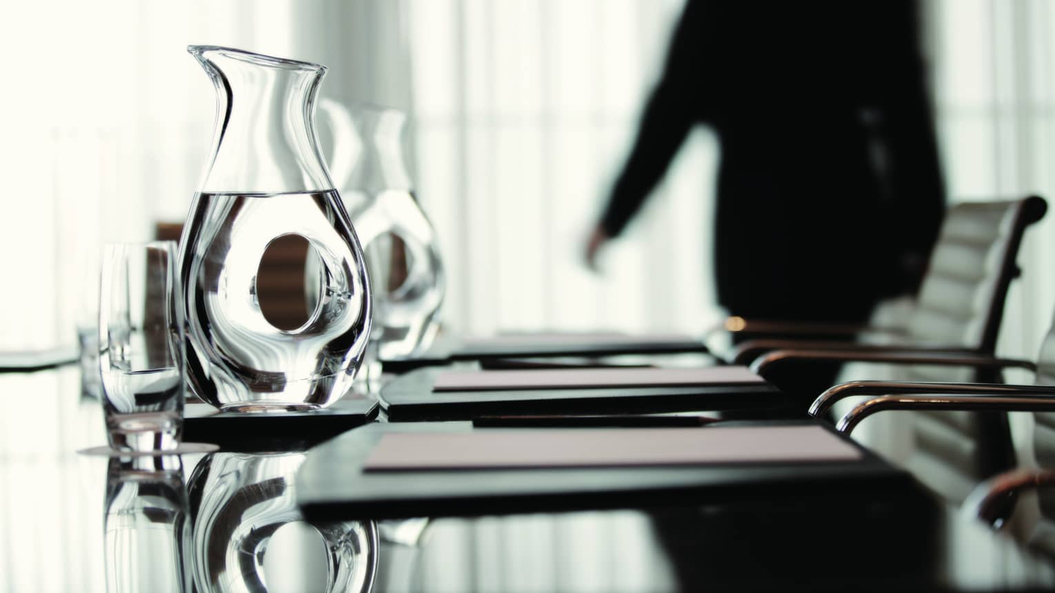 Person wearing business suit walks behind meeting table with agendas, glass water jugs