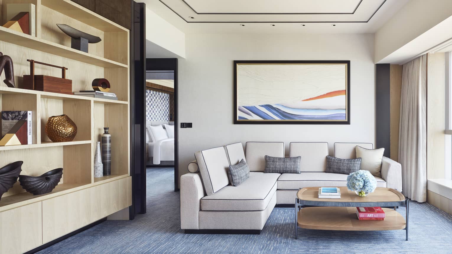 Living room with off-white, l-shaped sofa, blue carpet, wooden coffee table, large horizontal artwork, wall of shelves
