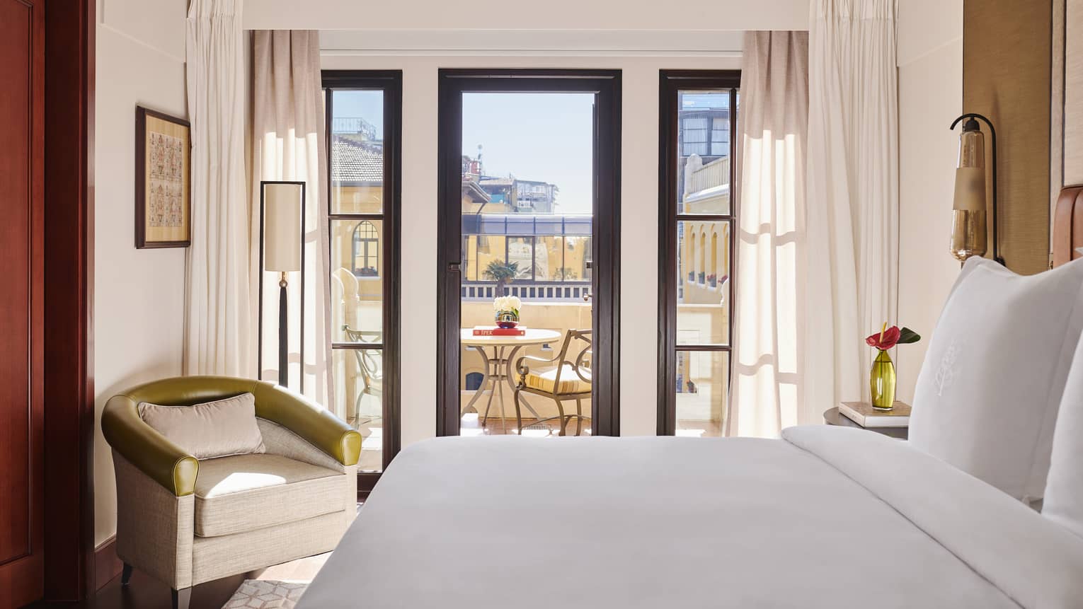 White bed against wall, beige club chair in corner and terrace view out wood-trimmed windows