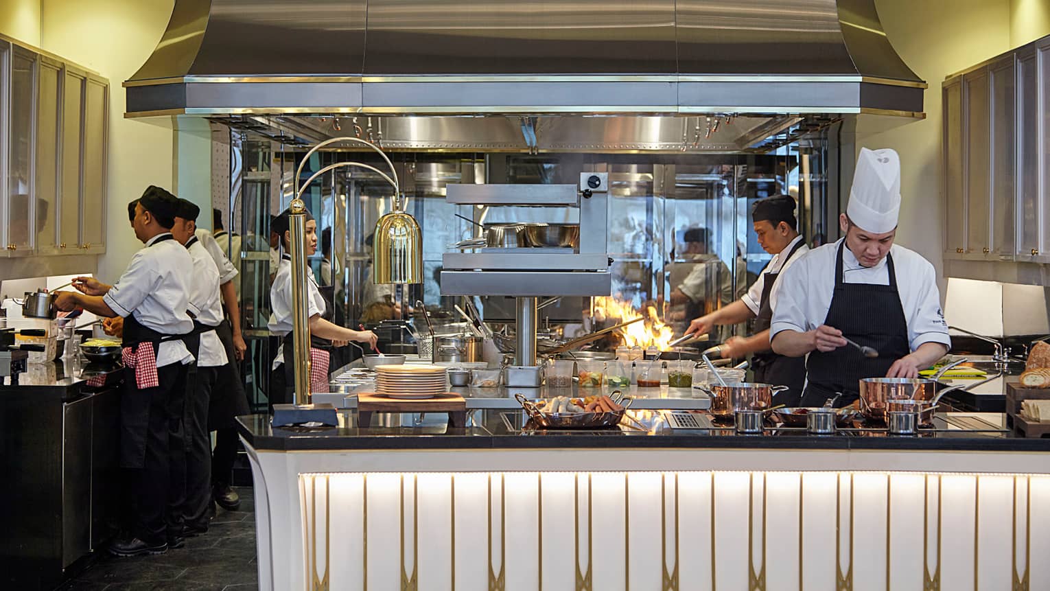 Chefs work on kitchen lines, flames shoot from pan in background