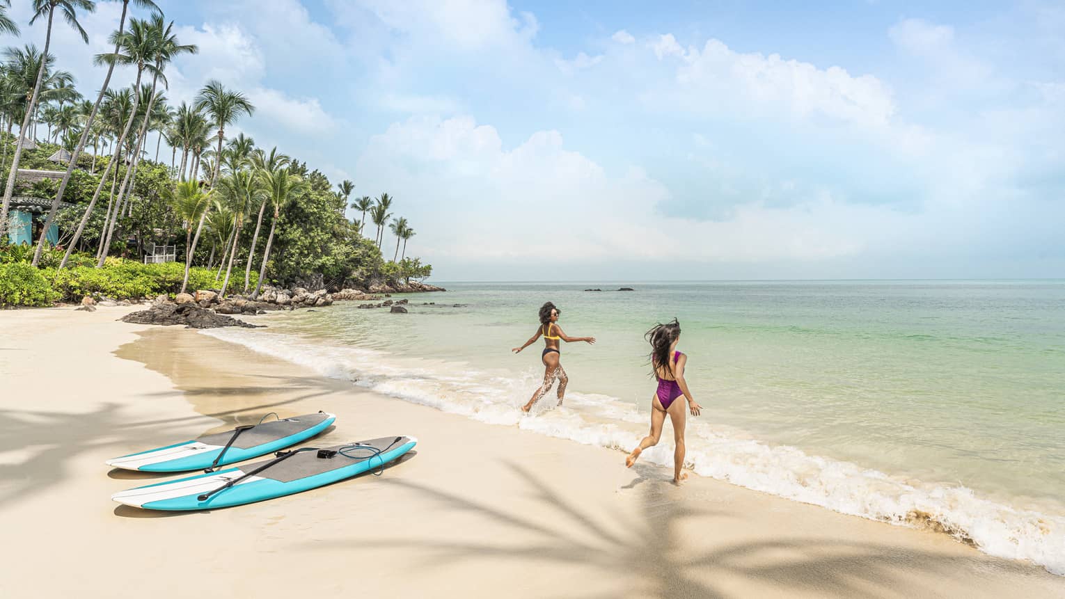 Two women in swimsuits wade into the water at the beach, their surfboards on the shore behind them