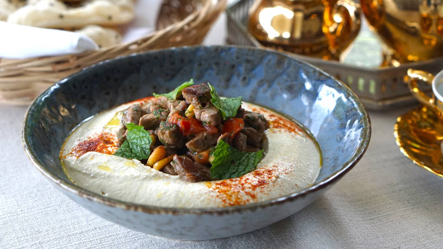 Blue bowl of hummus topped with za?atar-spiced lamb cubes, pine nuts, fresh mint leaves and paprika