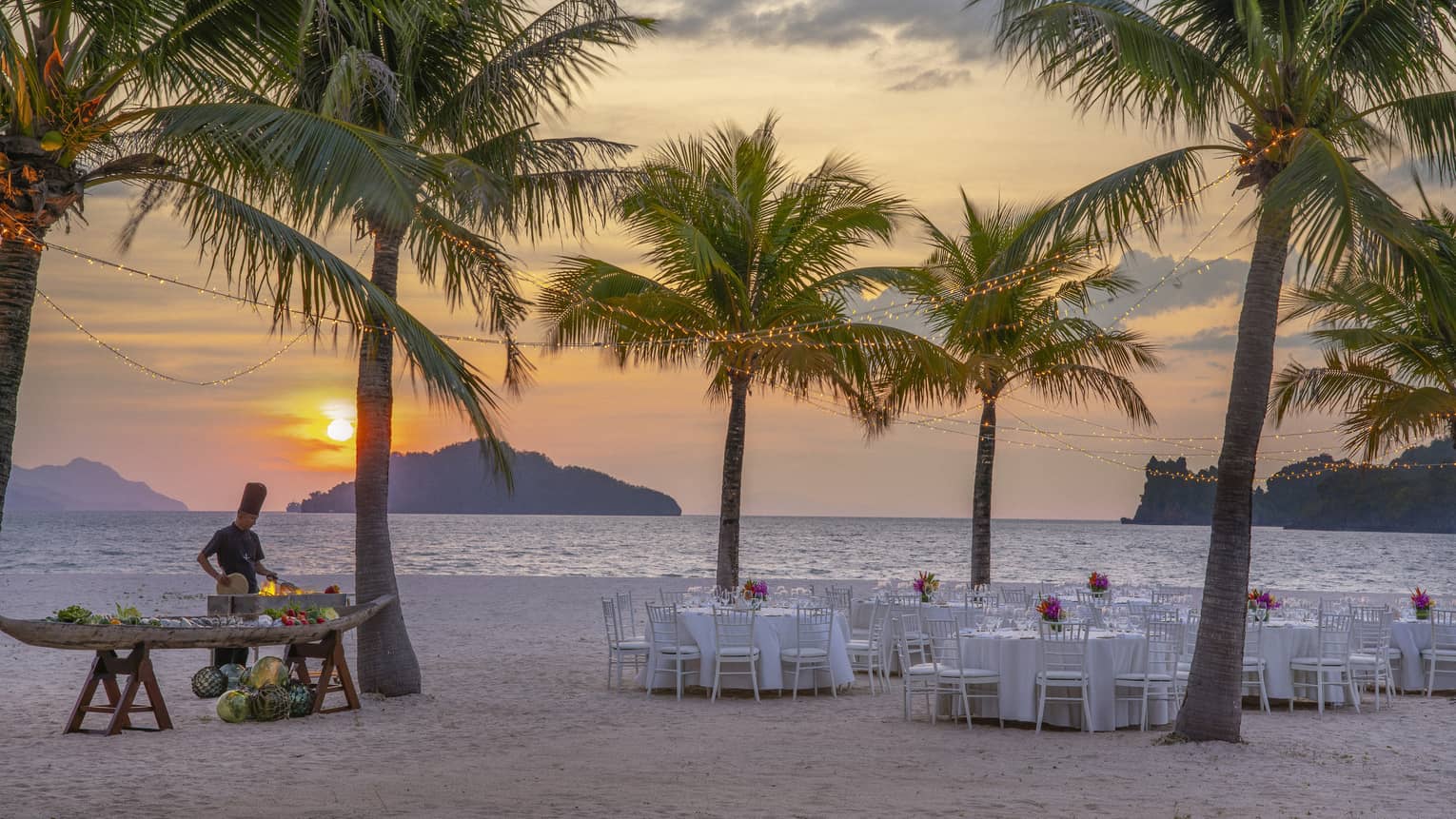 Tables set for dinner on the beach under a canopy of palm trees, a chef cooking over an open flame against the setting sun.