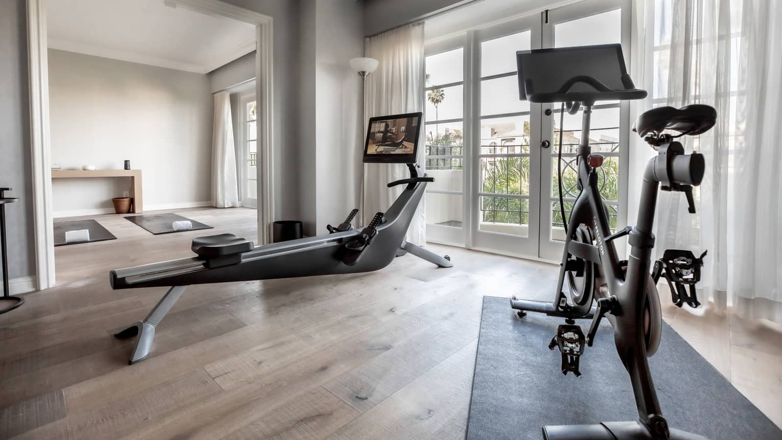 A hotel room with a rower and stationary bike facing window, wooden floors