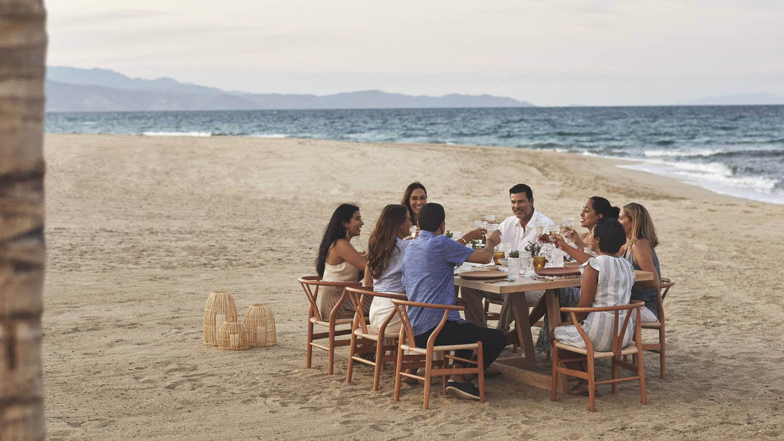 A group dines on the beach in wooden mid century modern dining chairs pulled up around a rectangular wooden dining table