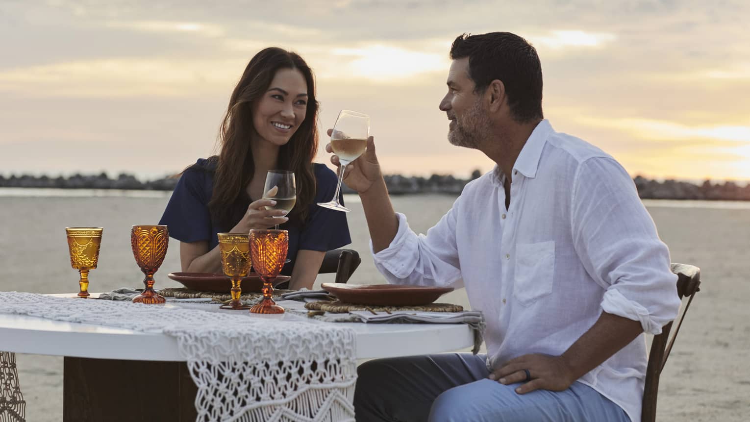 A smiling couple drinking wine against a sunset-streaked sky, at a beachfront dinner table set with amber-coloured goblets.