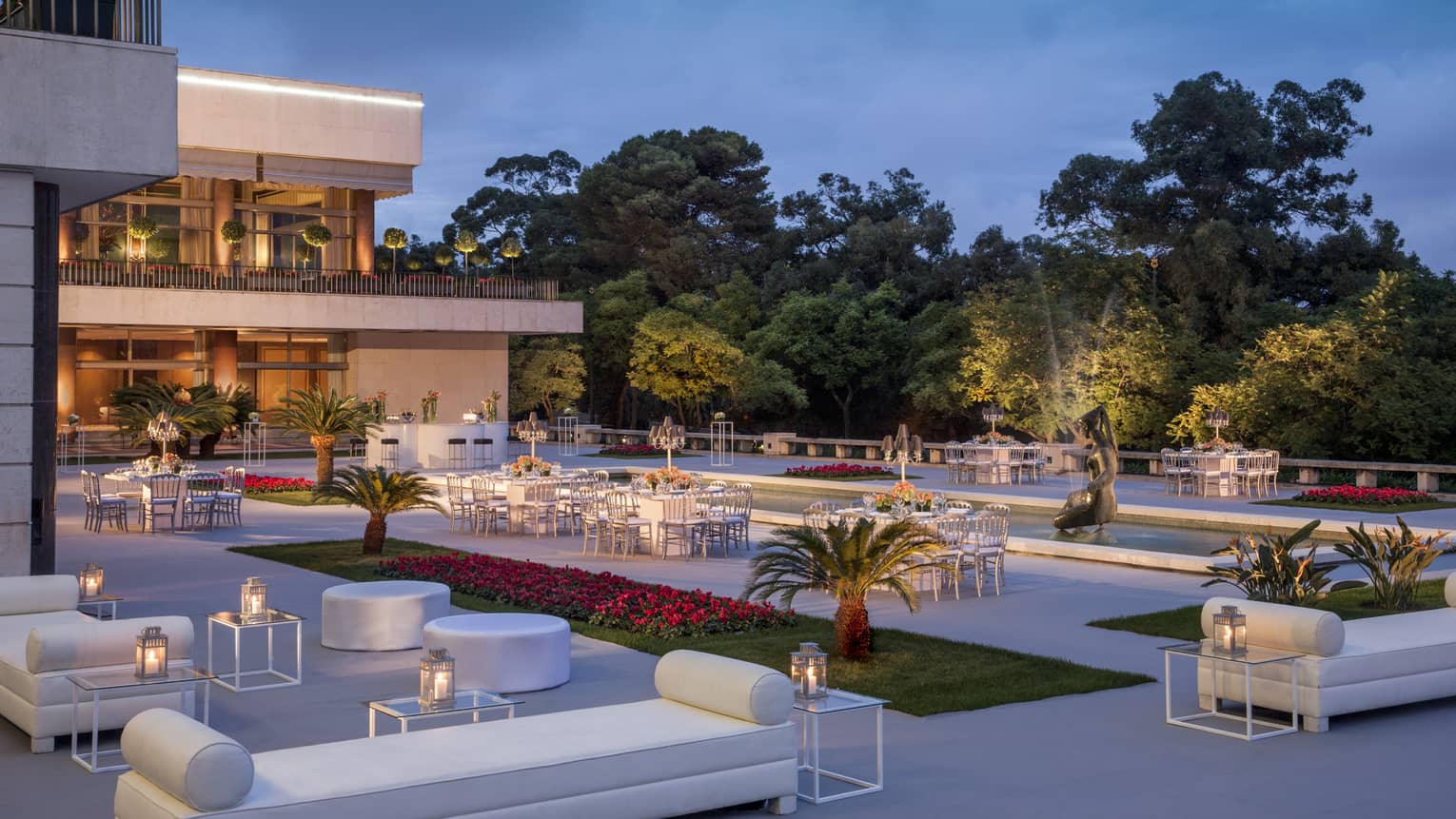 Modern white patio lounge furniture, tables, potted palms surrounding fountain at dusk