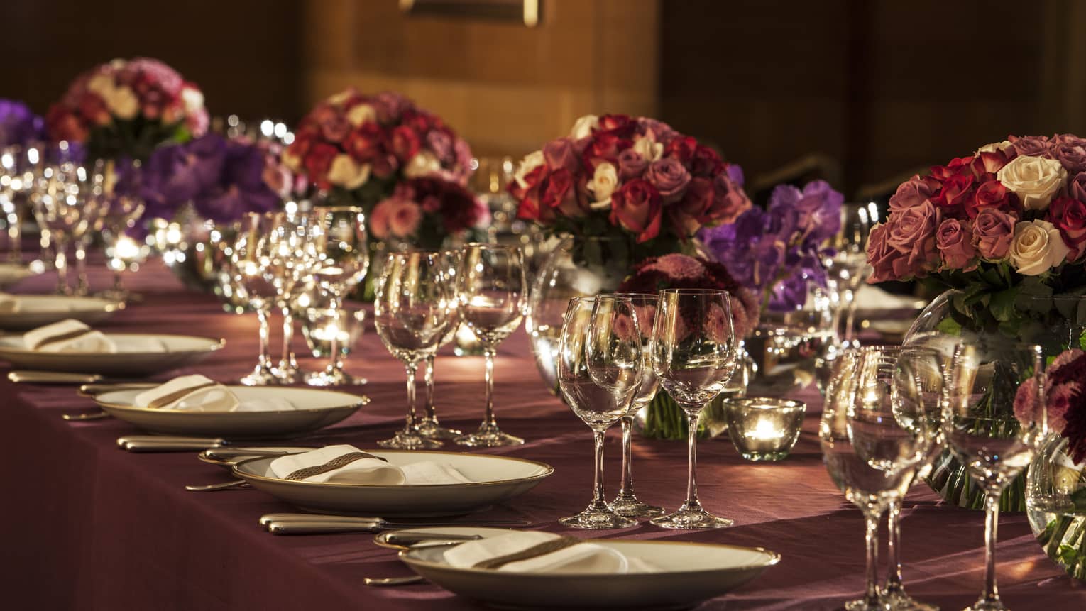 Deep pink and purple roses, wine glasses on elegant, candle-lit banquet table