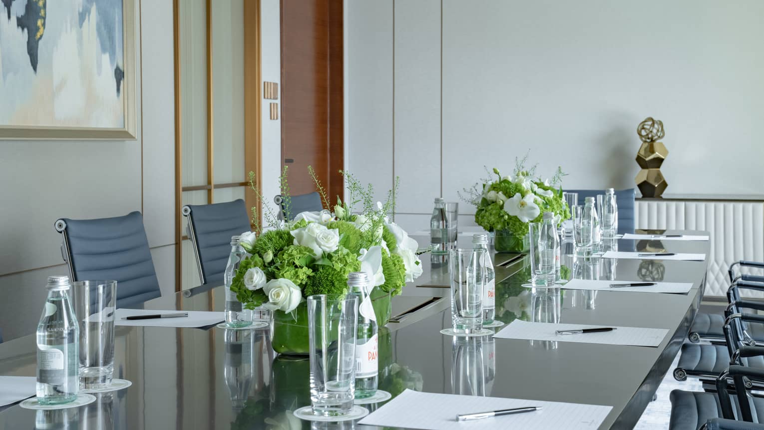 Meeting room with long boardroom table set with papers and pens, floral arrangements, bottles of water