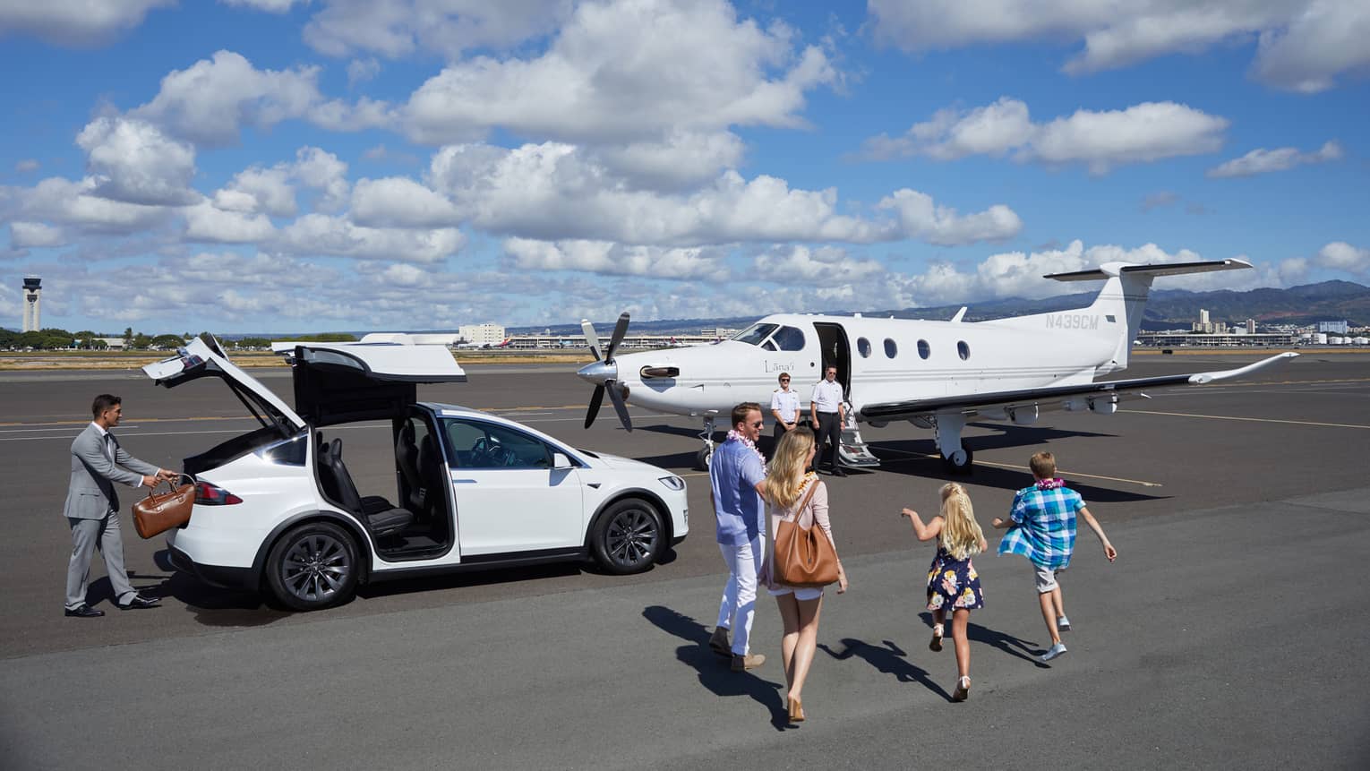 A family of four walks toward private plane while a man takes a suitcase out of a white car