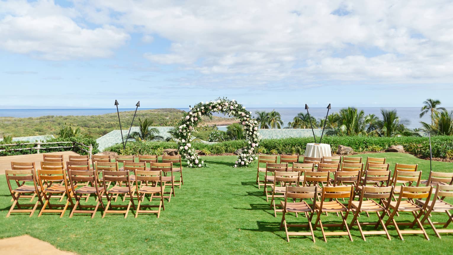 A series of wooden chairs set up in front of a floral arch and tiki torches, all overlooking the Lanai golf course.