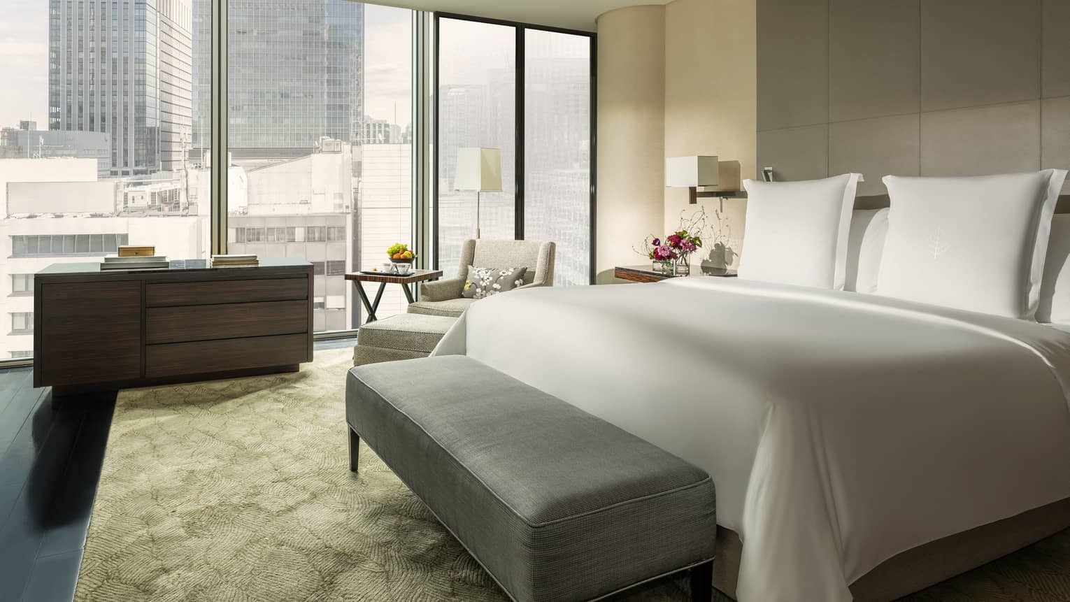 Hotel room with king bed, grey cushioned bench, dresser and wall of windows