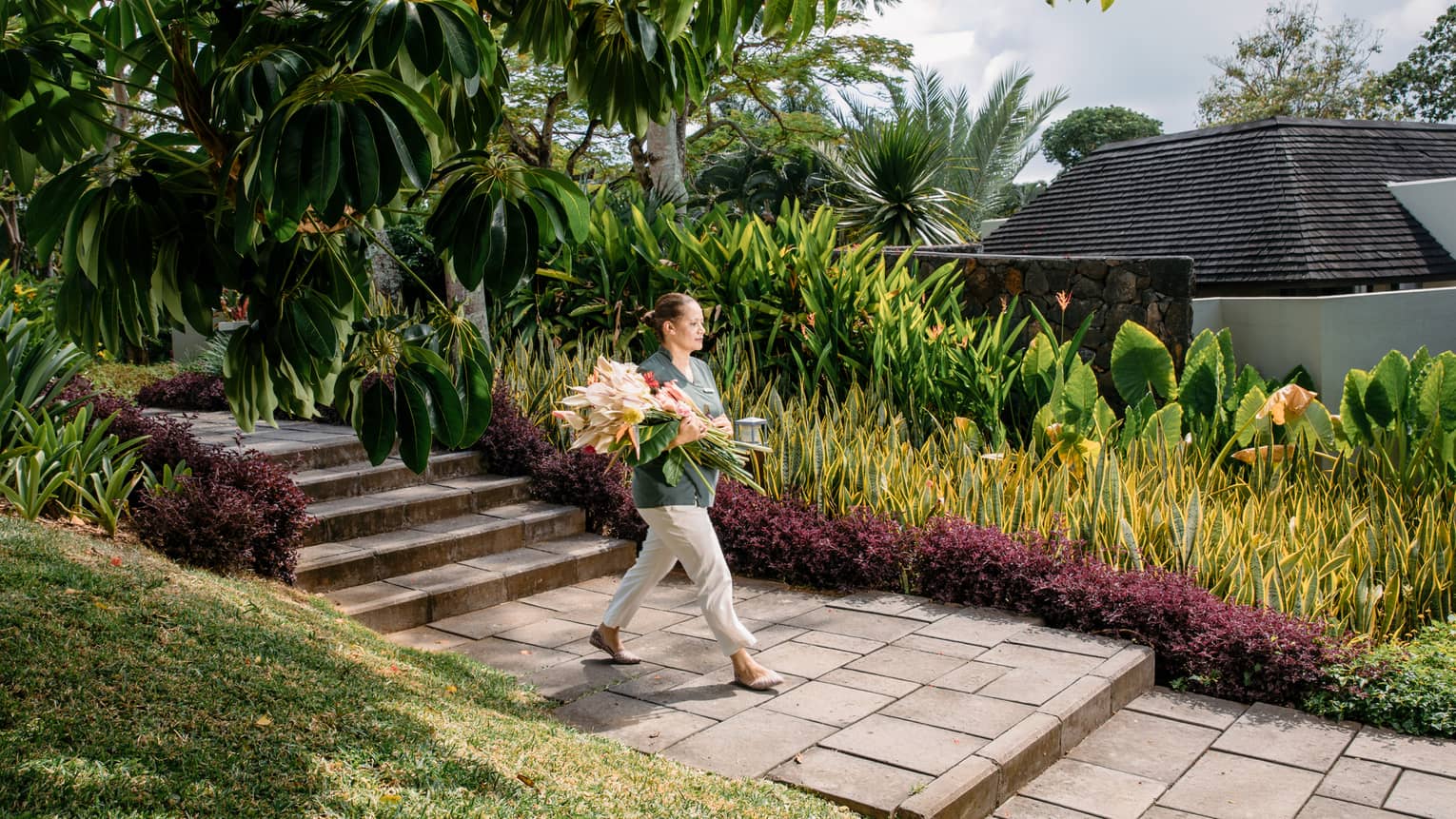 Hotel staff carries large bouquet of tropical flowers down brick path in garden