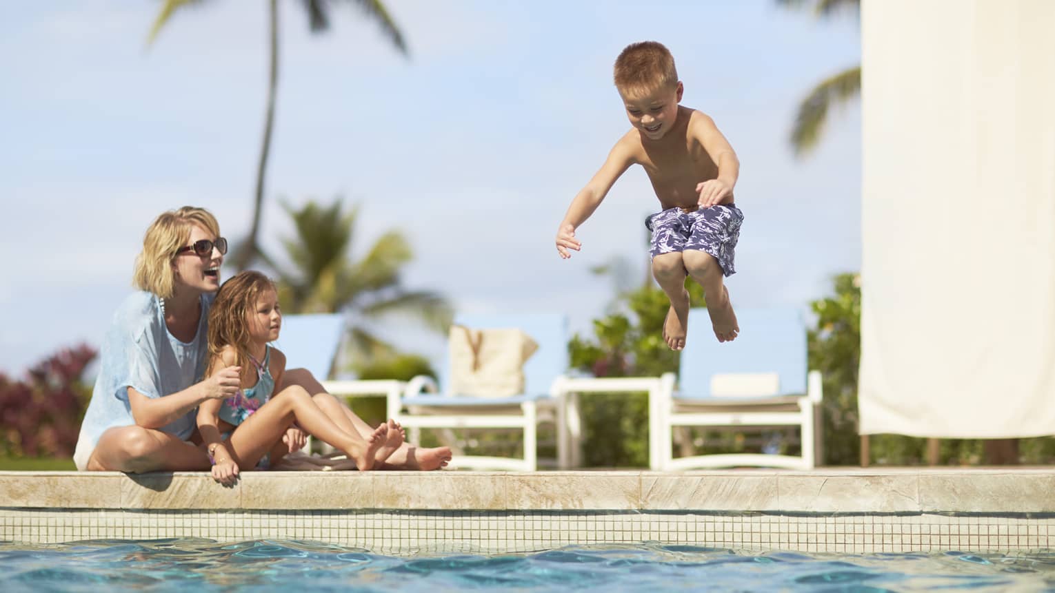 A small boy jumping into a pool next to his mom and little sister.