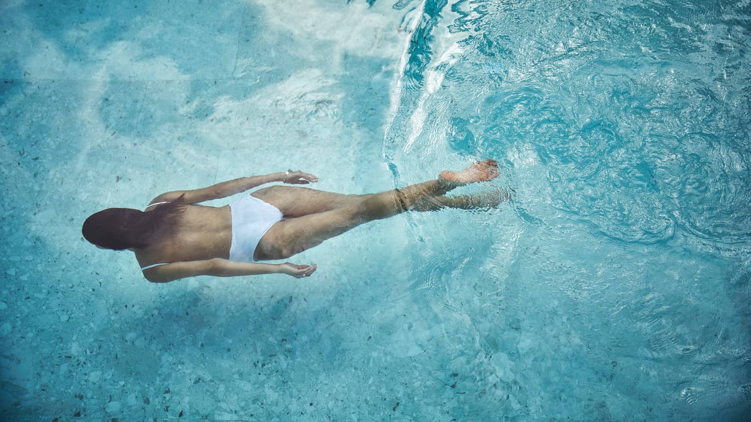 Woman in white swimsuit swimming underwater in blue pool