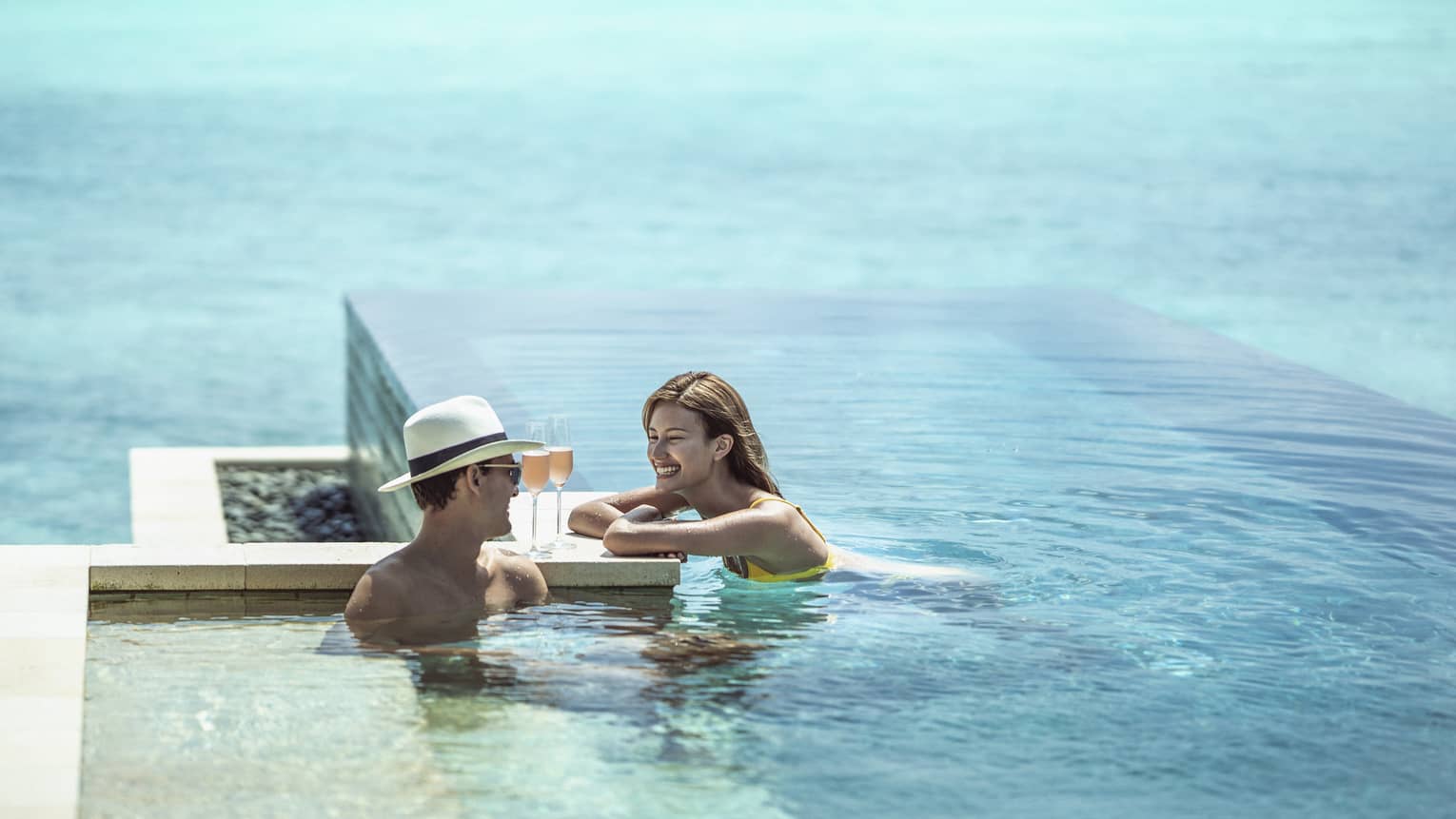 Smiling couple relaxes in infinity pool with glasses of champagne