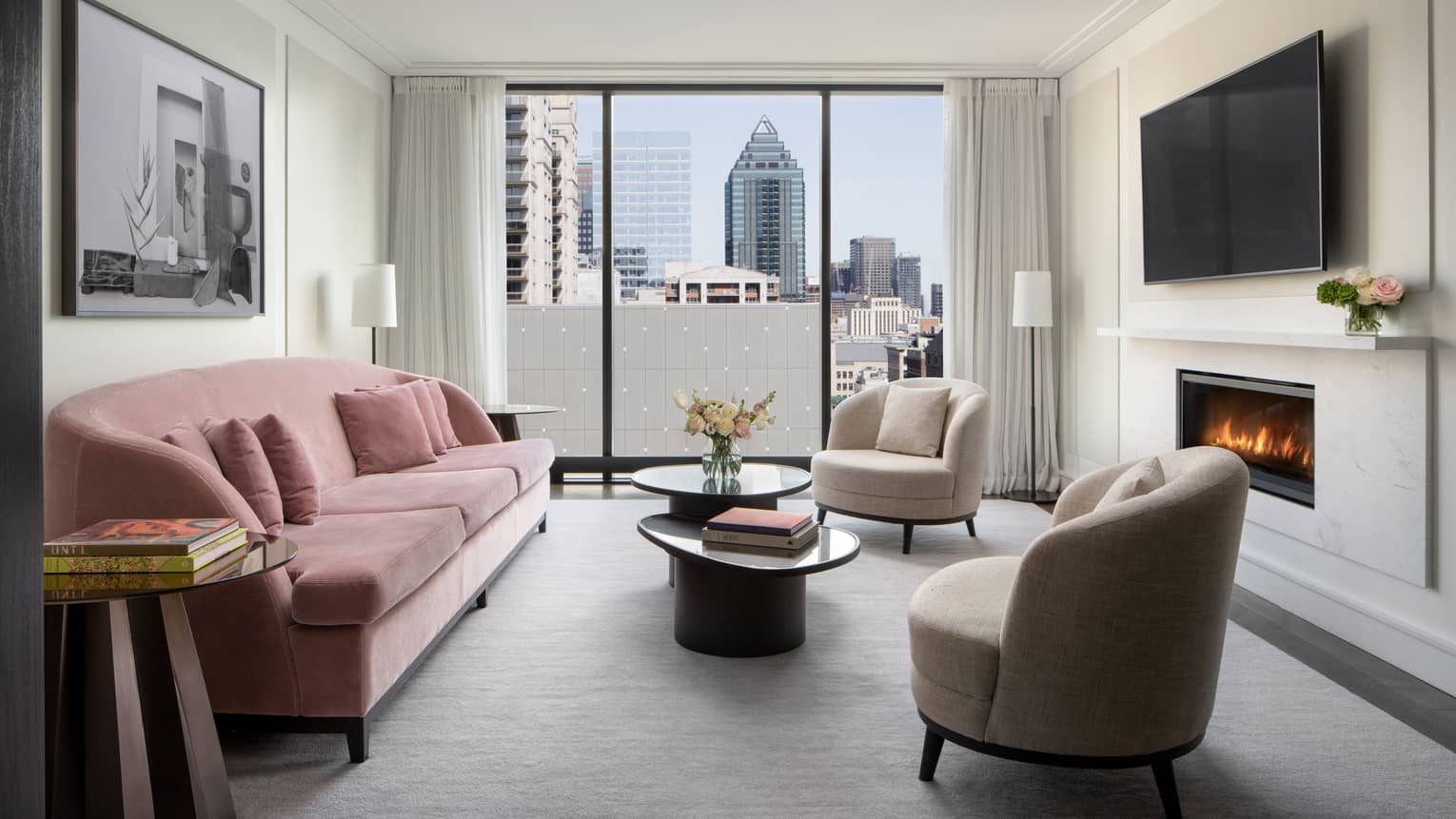 Hotel living room with pink velvet sofa, two arm chairs, fireplace, TV and city view