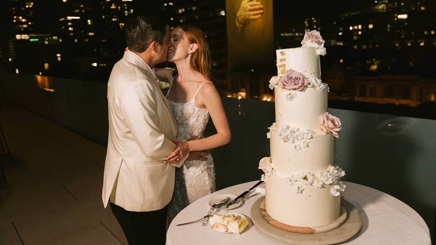 Woman wearing white sleeveless wedding dress and man wearing white tuxedo coat and black pants kiss on a rooftop terrace next to a wedding cake