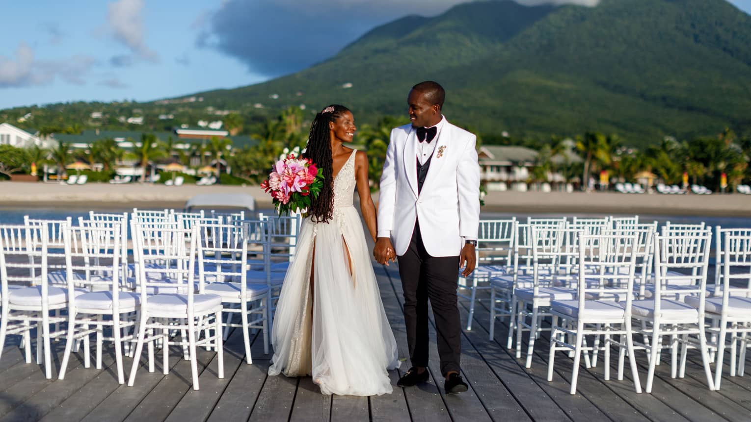 Woman in wedding dress, pink bouquet, holding hand of man in white jacket, black pants, chairs surrounding